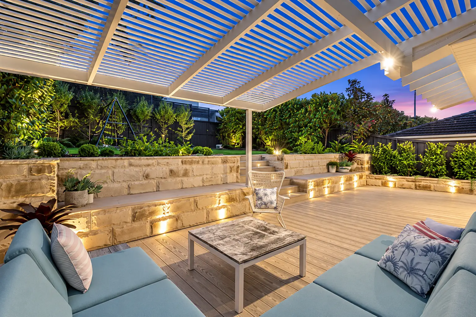 Timber deck beneath a louvred pergola with sandstone seat walls and garden lighting at dusk