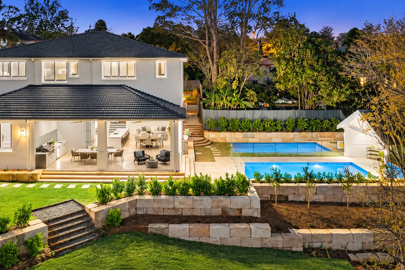 Turramurra garden at dusk, with a lit pool, glass fencing, tiered sandstone retaining walls and a covered alfresco beside the house