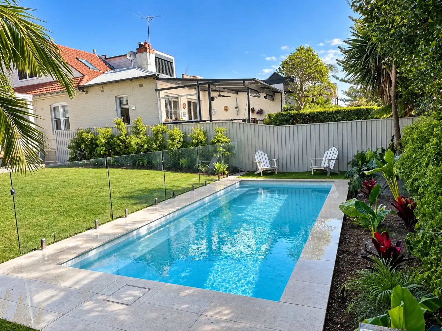 Sunny rectangular swimming pool with pale stone paving, glass fencing, lawn and tropical planting in Mosman