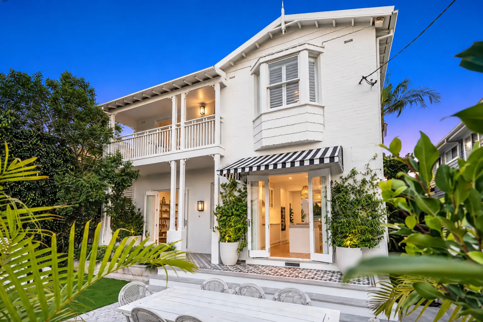 White weatherboard home at dusk with a black and white striped awning, patterned tile terrace, potted bays and outdoor dining