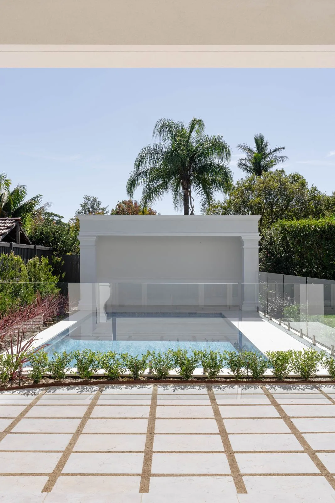 Pale stone pavers with gravel joints leading to a glass-fenced pool and cabana