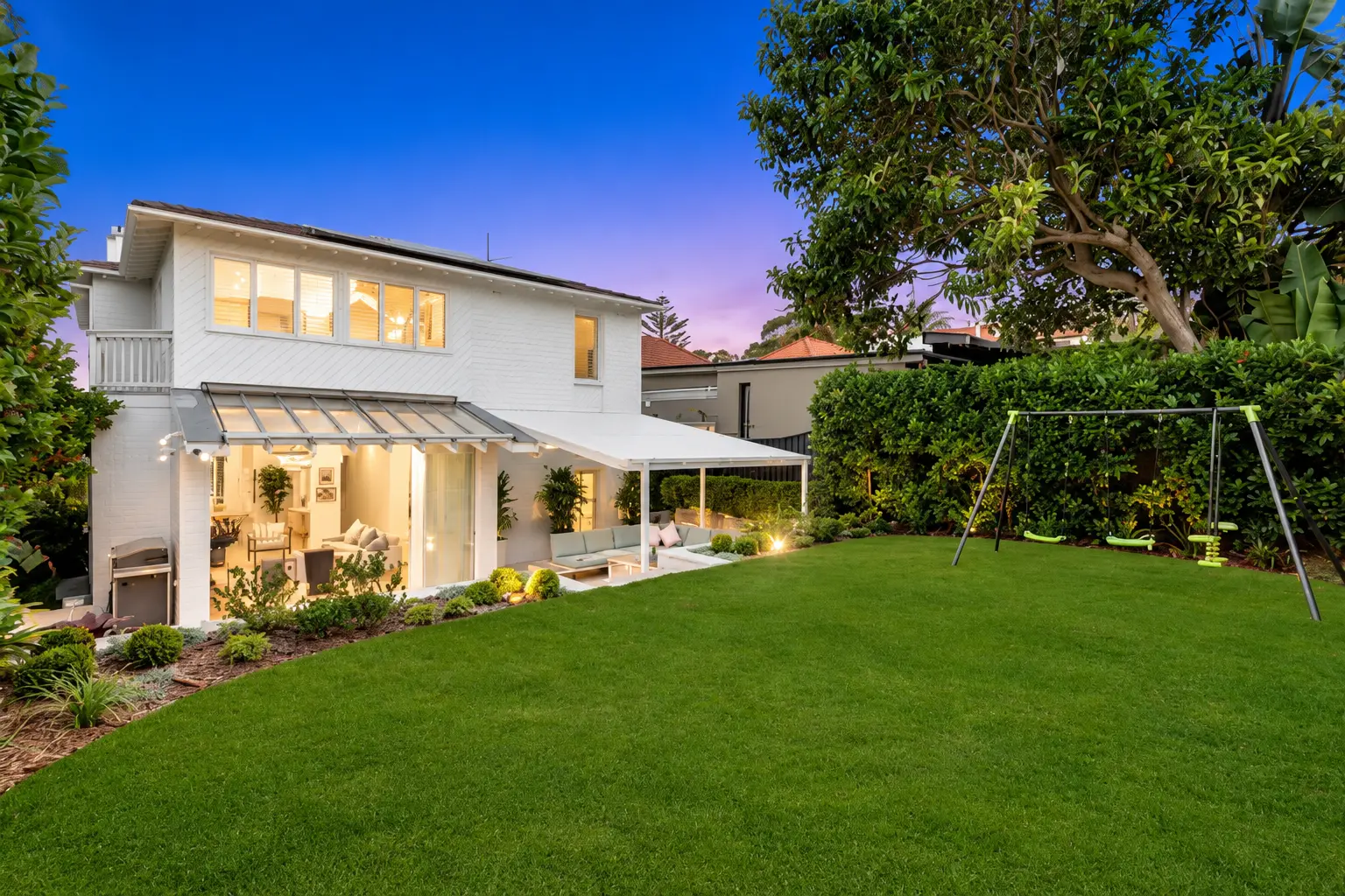 Level lawn with a swing set beside a white weatherboard home and layered garden beds at dusk