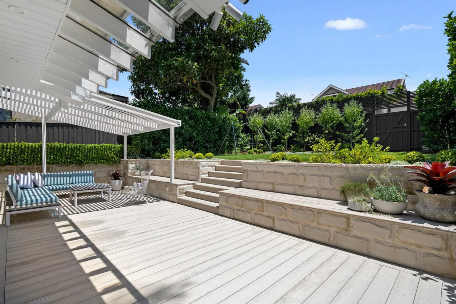 Timber deck with sandstone steps up to a lawn, swing set, hedging and planters on a sunny day